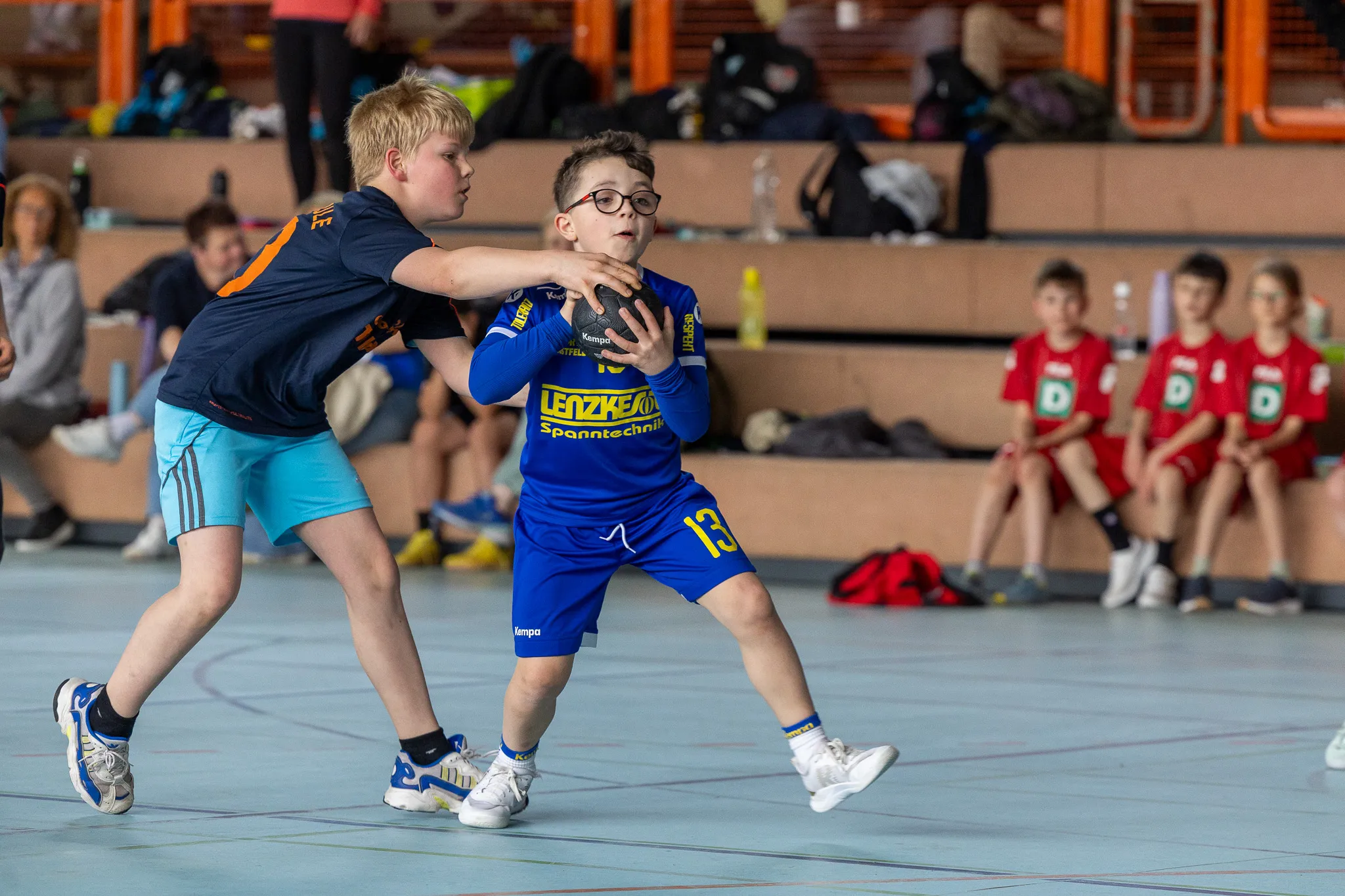 Zwei junge Handballspieler im Spiel: Ein Spieler in blauem Trikot mit Brille versucht, den Ball zu halten, während ein Gegner in dunklem Trikot ihn blockt.