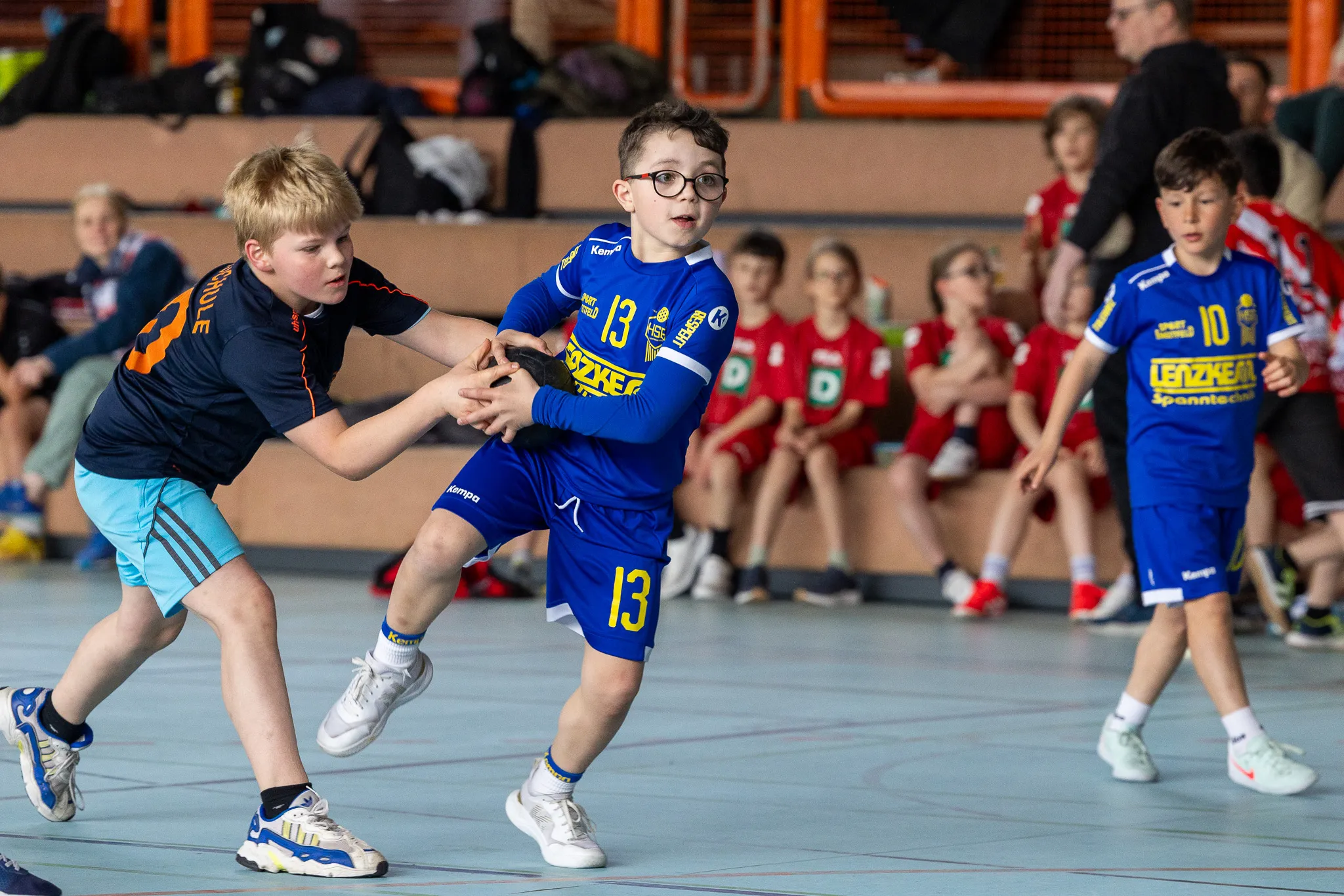Zwei junge Handballspieler kämpfen um den Ball, während im Hintergrund weitere Spieler zusehen.