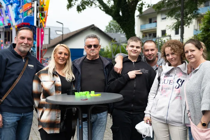 Eine Gruppe von sieben Menschen steht lächelnd vor einer Kirmes. Im Hintergrund sind Fahrgeschäfte und Buden zu sehen.