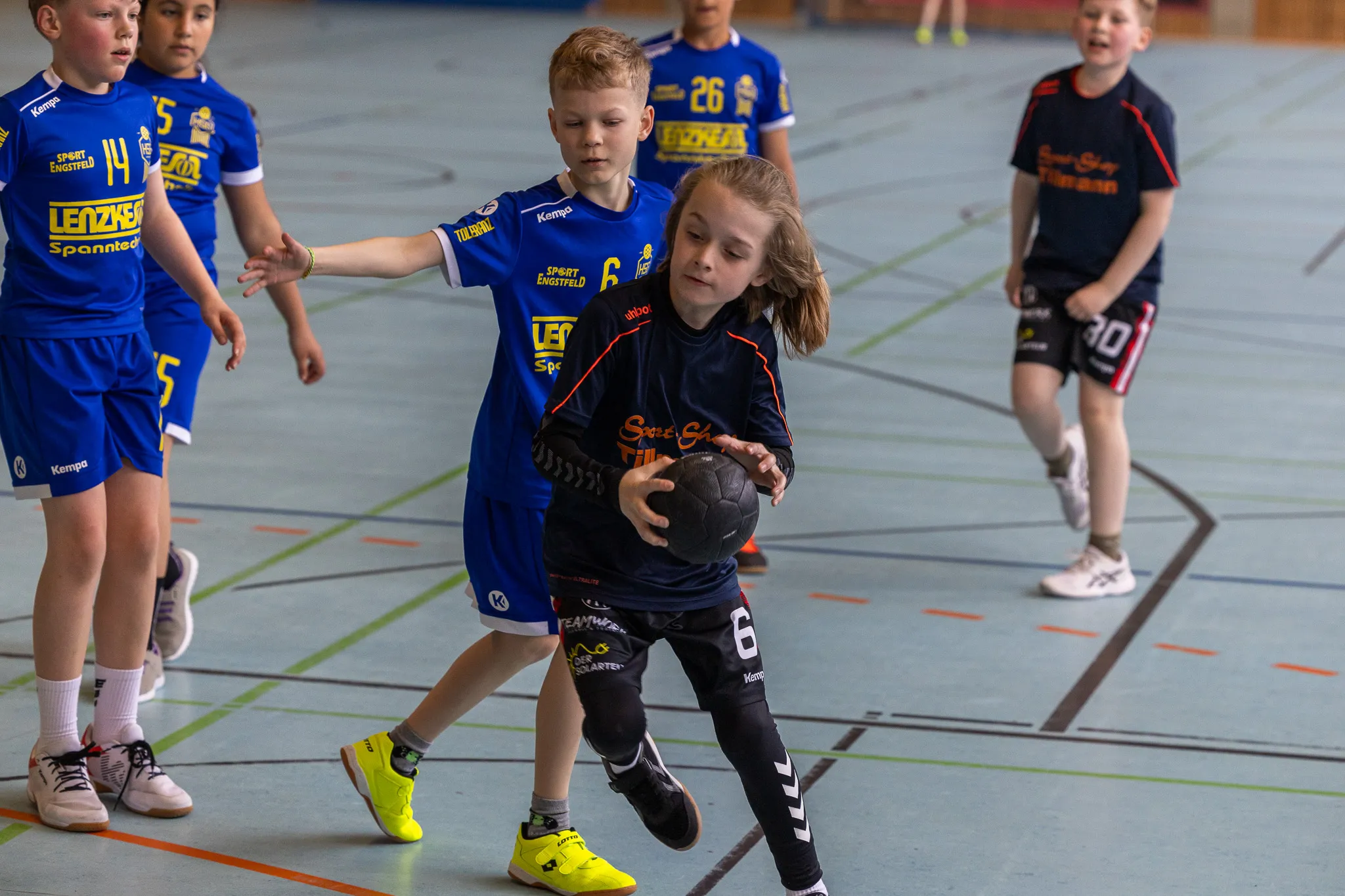 Zwei junge Handballspieler kämpfen um den Ball in einer Sporthalle. Der Spieler im blauen Trikot blockiert die Spielerin mit rotem Trikot und gelben Schuhen.