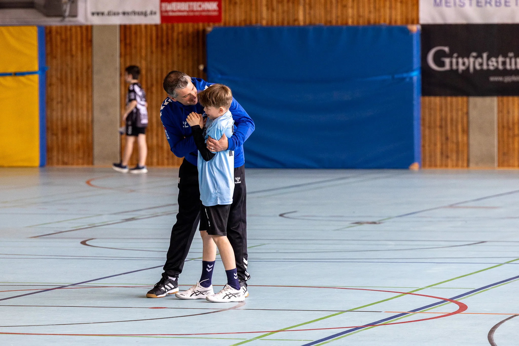 Ein Mann umarmt einen Jungen in Sportkleidung in einer Turnhalle. Im Hintergrund sind weitere Personen und Werbebanner zu sehen.