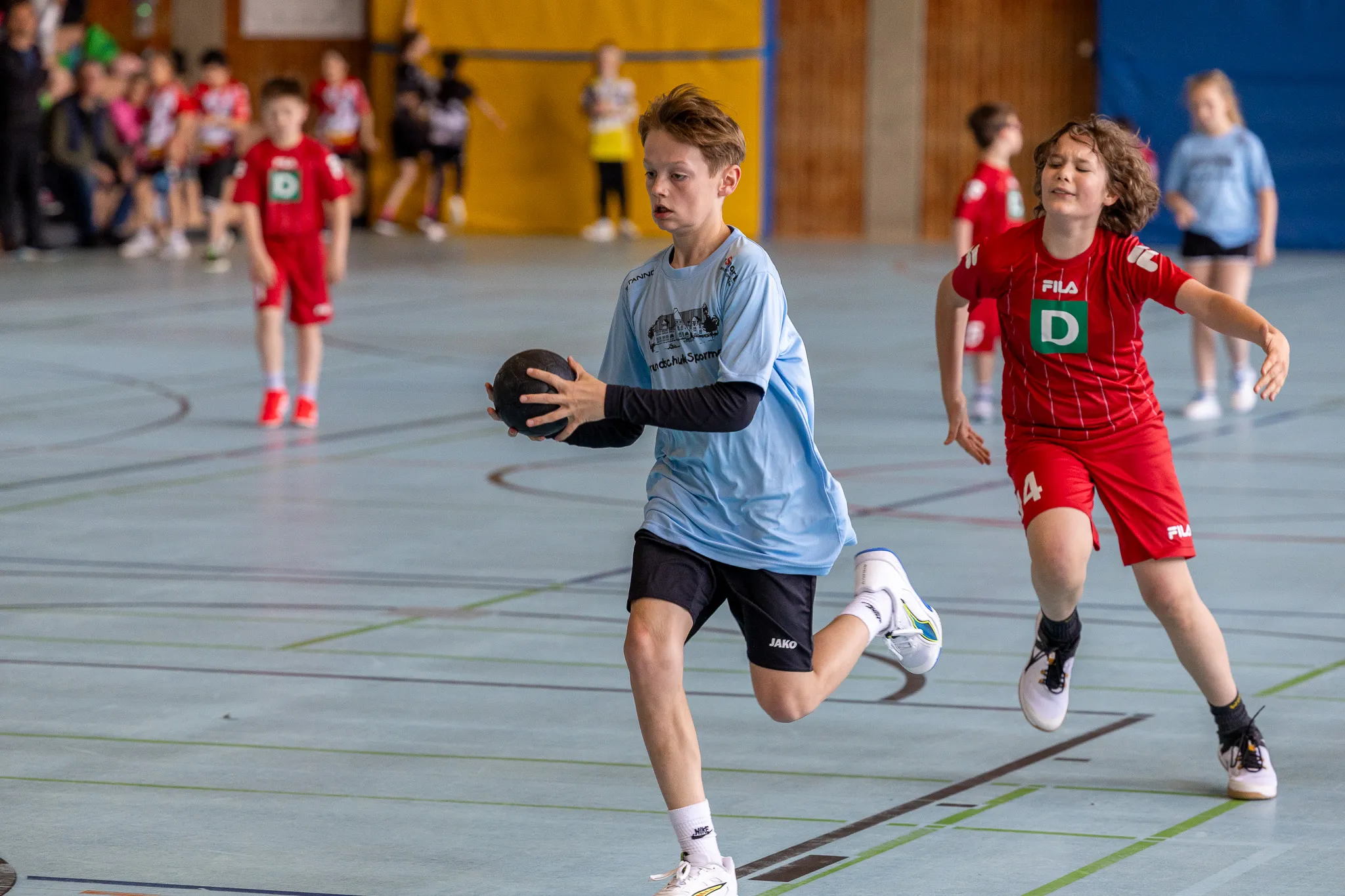 Ein Jugendlicher mit Ball läuft beim Handballspiel in einer Sporthalle, während ein Gegenspieler ihn verfolgt. Im Hintergrund sind weitere Spieler und Zuschauer zu sehen.