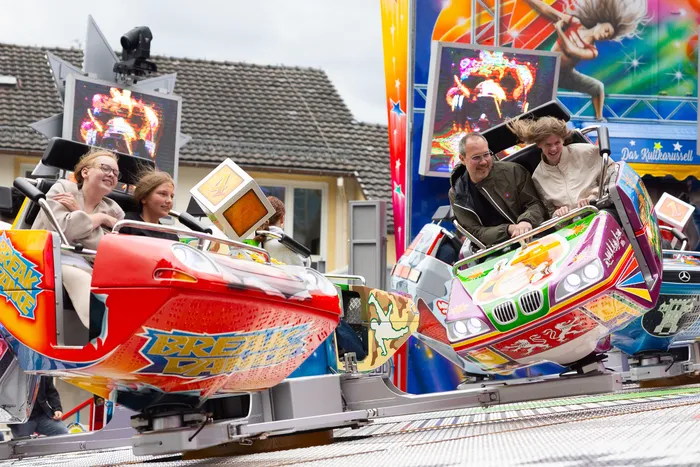 Zwei rote Fahrgeschäfte im Stil von Autos drehen sich auf der Kirmes, besetzt mit Familien mit Kindern. Im Hintergrund ist ein buntes Karussell zu sehen.