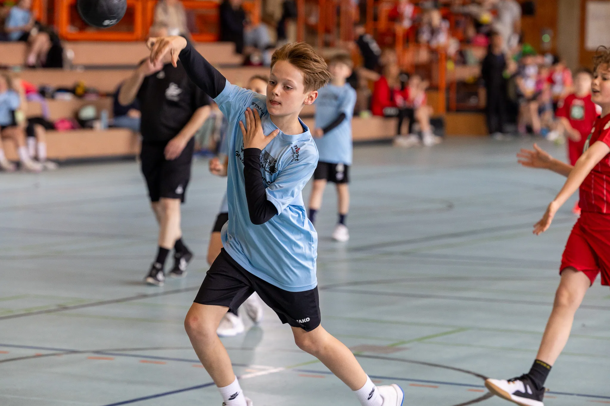 Ein Jugendlicher wirft einen Handball in einer Sporthalle. Im Hintergrund sind weitere Spieler und Zuschauer zu sehen.