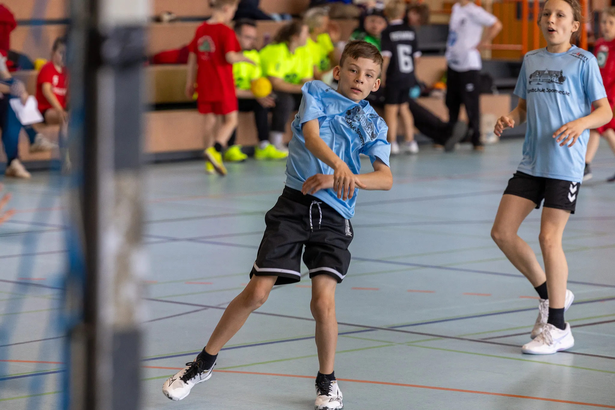 Zwei Jungen beim Handballspiel in einer Sporthalle. Der Junge im Vordergrund springt zum Wurf ab, während sein Teamkollege im Hintergrund bereit steht.