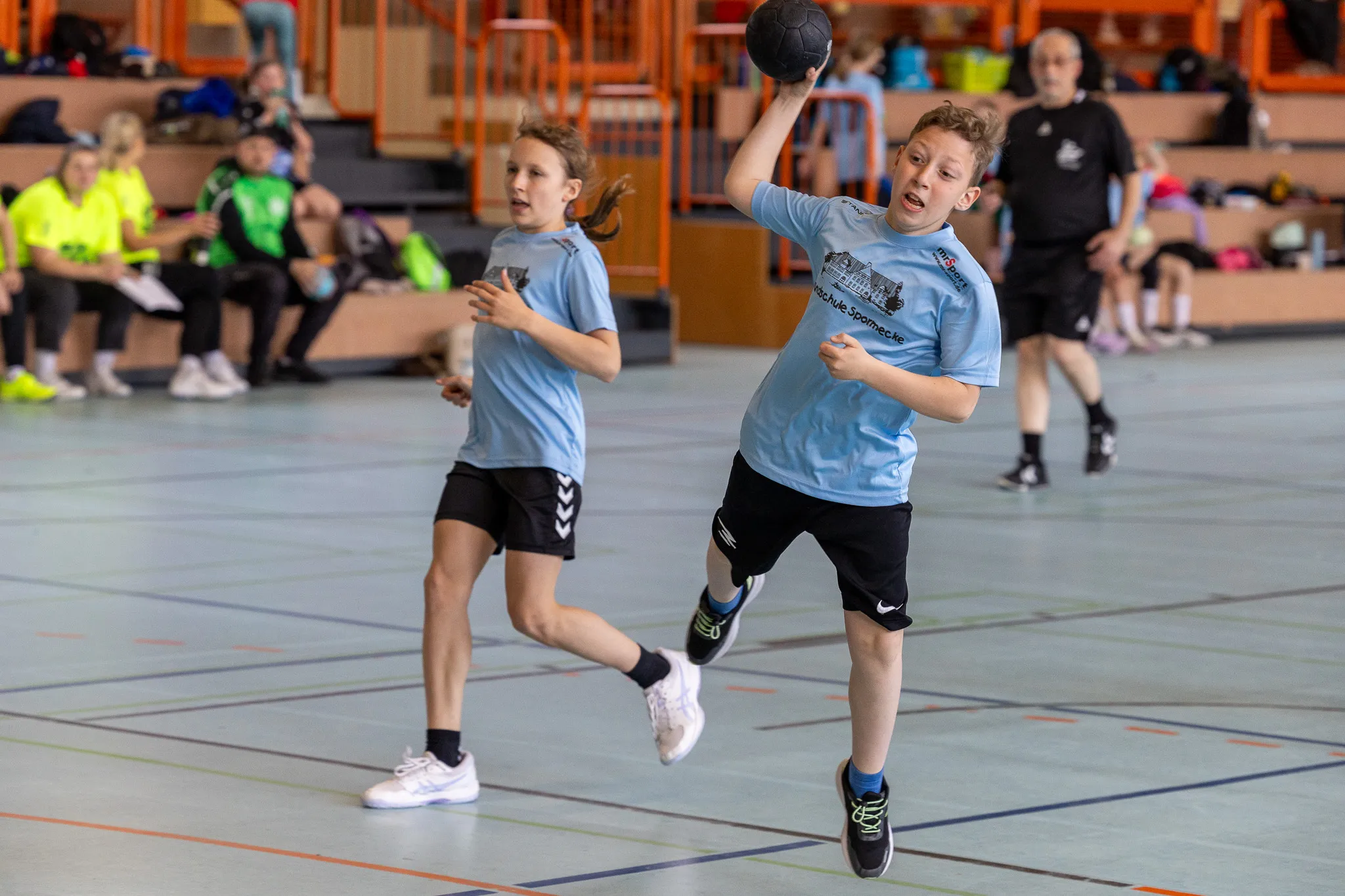 Zwei junge Handballspielerinnen in blauen Trikots trainieren in einer Sporthalle. Die Spielerin rechts wirft den Ball, während ihre Teamkollegin bereit für den Pass ist.
