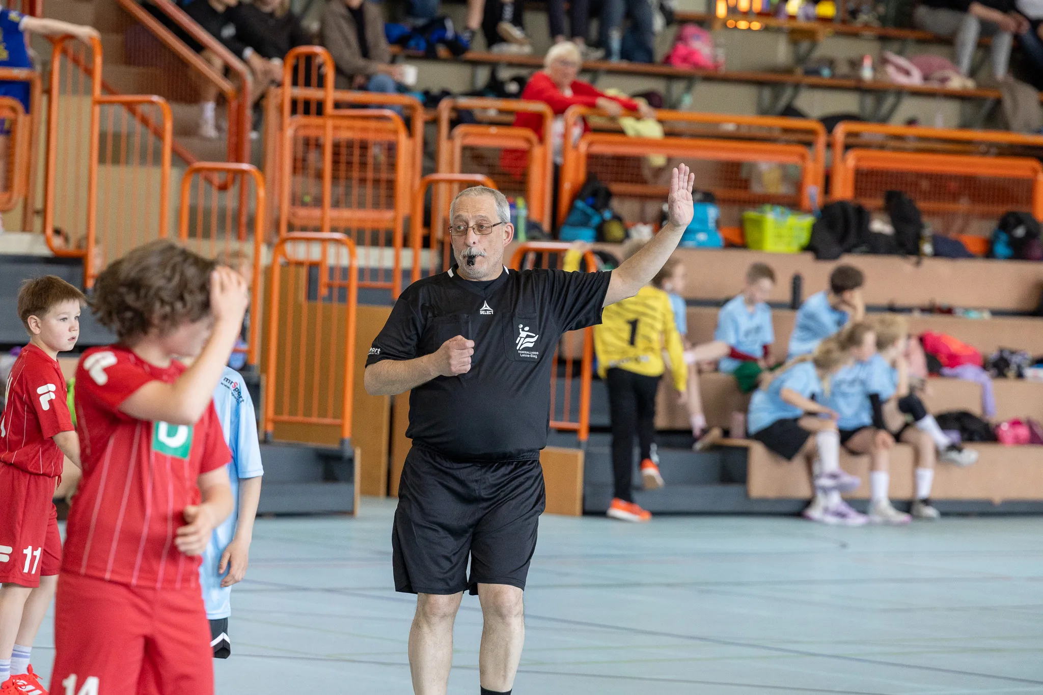 Handballtrainer mit erhobener Hand gestikuliert während eines Spiels von Kindern in roten Trikots in einer Sporthalle.