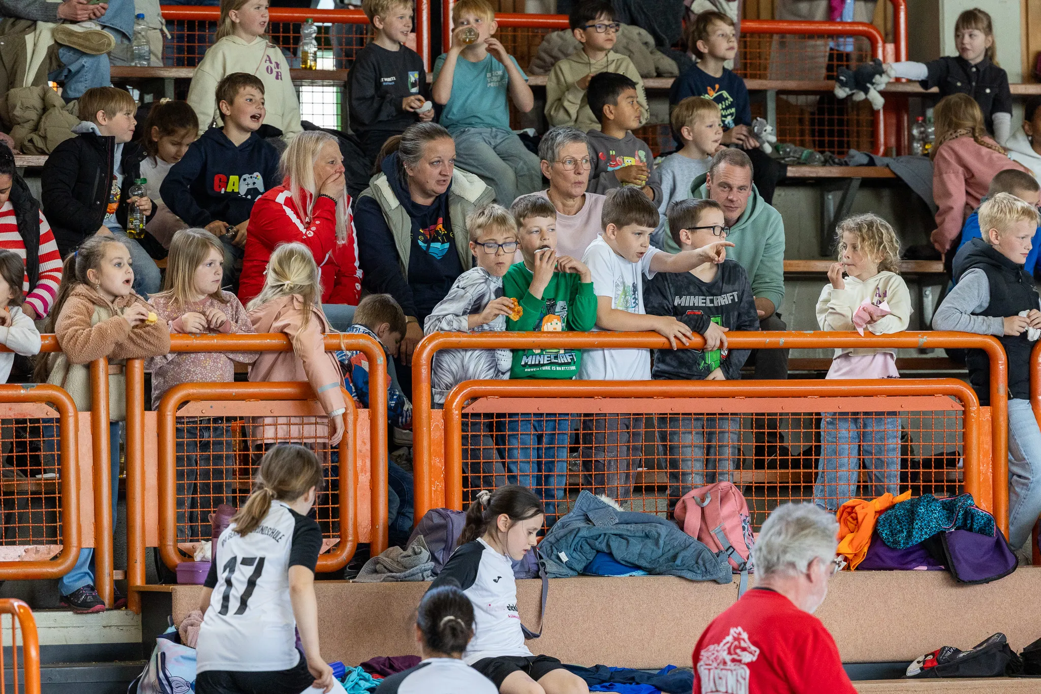 Zuschauer, darunter viele Kinder, sitzen auf Tribünen in einer Sporthalle und verfolgen ein Spiel. Im Vordergrund sitzen zwei junge Volleyballspielerinnen auf einer Bank.