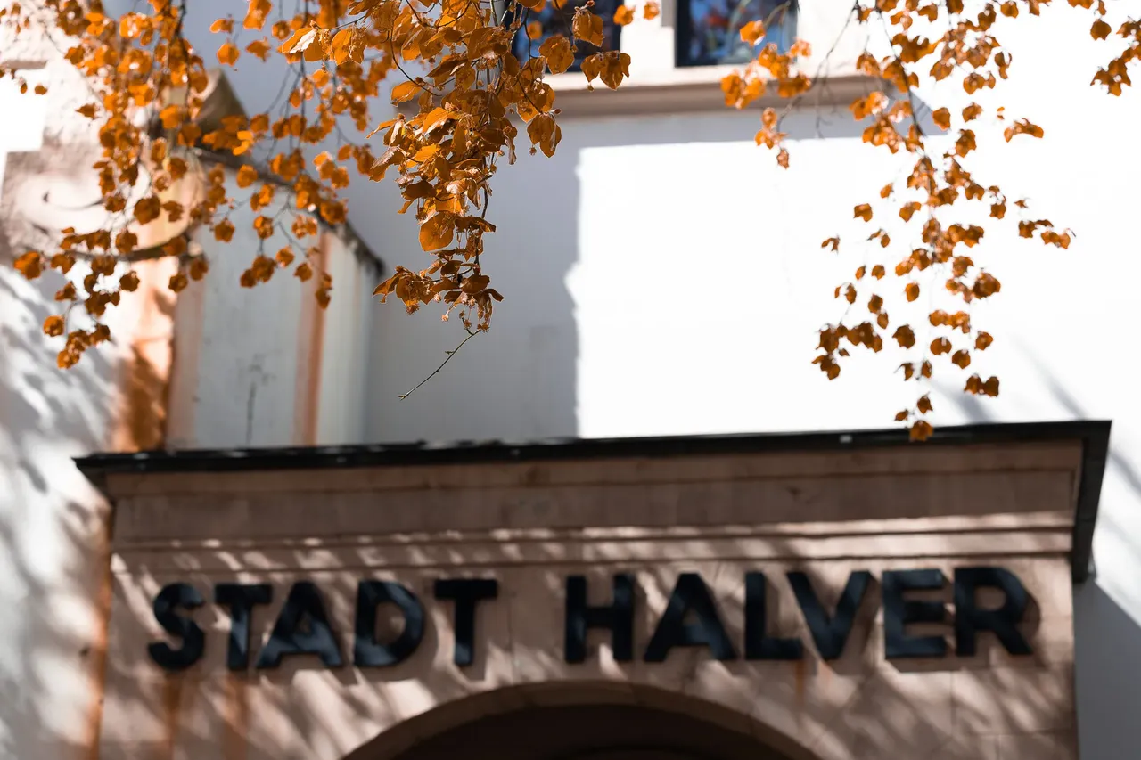 Schild mit der Aufschrift „STADT HALVER“ an einem hellen Gebäude, Äste mit Blättern im oberen Bildbereich.