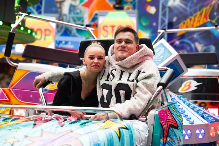 Ein junges Paar sitzt lächelnd in einer Fahrgeschäftskabine auf der Kirmes, umgeben von bunten Lichtern und Dekorationen.