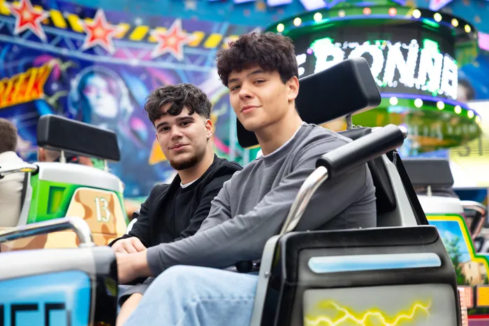 Zwei junge Männer sitzen auf einem Fahrgeschäft auf der Kirmes und schauen in die Kamera.