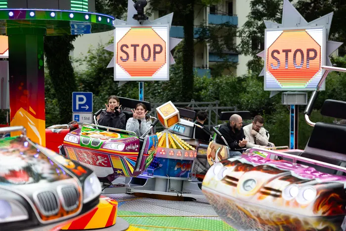 Fahrende Menschen in bunt dekorierten Autos auf einer Karussell-Fahrt auf der Kirmes. Im Hintergrund sind weitere Fahrgeschäfte und ein Parkplatzschild zu sehen.