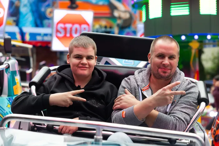 Zwei junge Männer sitzen auf einer Kirmesattraktion und machen mit den Händen das Victory-Zeichen. Im Hintergrund sind weitere Fahrgeschäfte und Lichter zu sehen.
