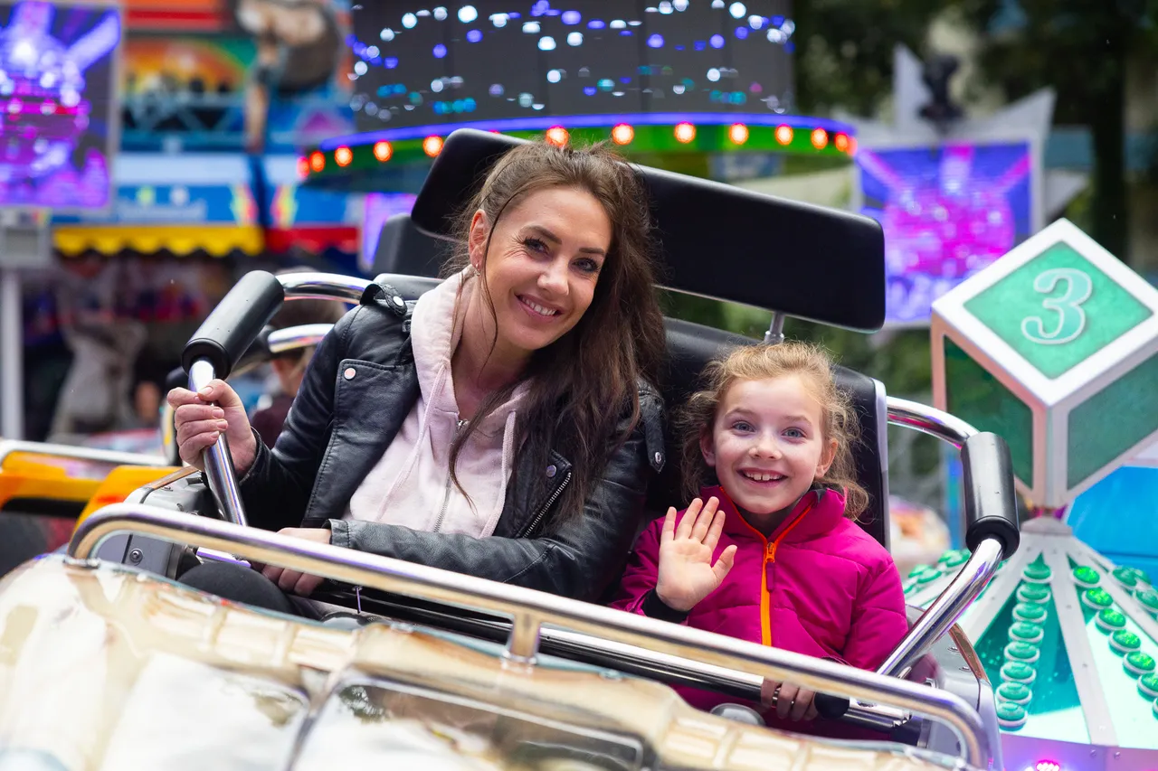 Eine Frau und ein kleines Mädchen sitzen lächelnd in einer Karussell-Fahrt und winken in die Kamera. Im Hintergrund sind weitere Fahrgeschäfte und Lichter einer Kirmes zu sehen.