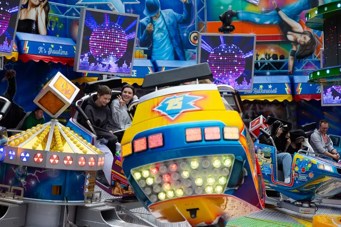 Zwei junge Frauen lächeln in einem bunt gestalteten Karussellfahrzeug, während sich die Attraktion dreht. Im Hintergrund sind weitere Fahrgeschäfte und Besucher der Kirmes zu sehen.