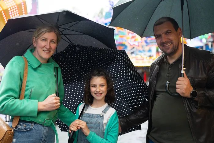 Familie mit Kind lächelt in die Kamera, geschützt unter Regenschirmen vor dem bunten Hintergrund der Kirmes.