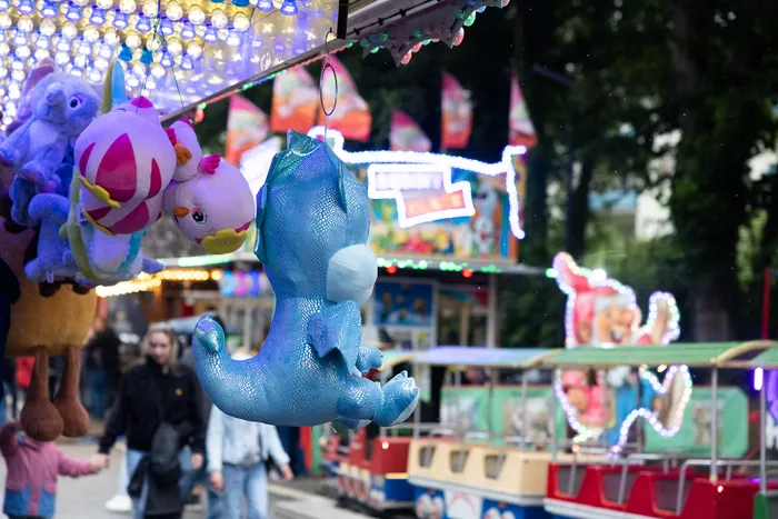 Ein blauer Plüschdinosaurier hängt als Gewinn an einem Kirmesstand vor Menschen und einem kleinen Zug.