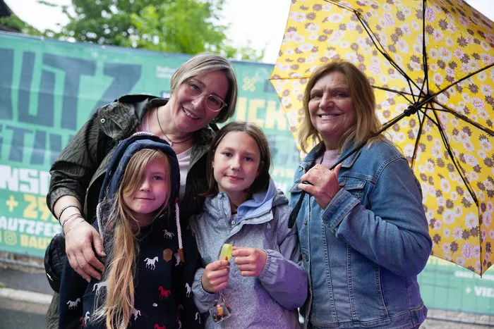 Vier Personen lächeln in die Kamera: zwei Frauen und zwei Kinder. Im Hintergrund ist ein buntes Werbeschild für die Kirmes zu sehen.
