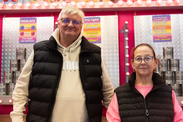 Zwei ältere Menschen stehen vor einem Jahrmarktstand mit Konservendosen. Der Mann trägt eine schwarze Jacke mit Kapuze und die Frau eine pinkfarbene Jacke.