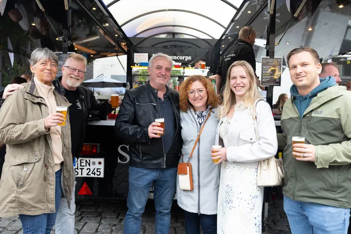 Sechs Menschen stehen lächelnd mit Biergläsern und einem Glas Orangensaft vor einem Getränkestand auf der Halveraner Kirmes.