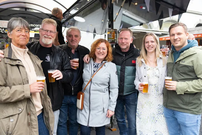 Eine Gruppe von sieben Menschen steht lächelnd zusammen und hält Biergläser in der Hand, im Hintergrund ein Kirmesstand.