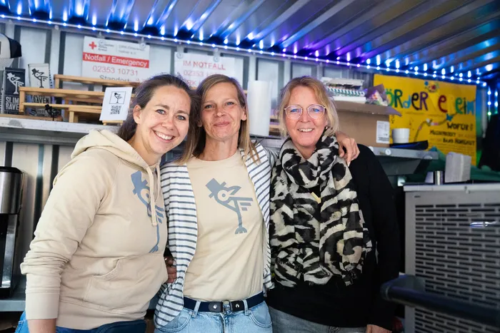 Drei lächelnde Frauen stehen eng zusammen vor einem Kirmesstand mit Notfallnummern und Getränkeangeboten.