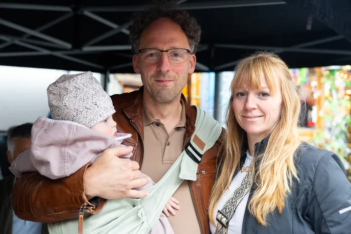 Ein Mann mit Baby in einem Tragetuch und eine Frau lächeln in die Kamera. Im Hintergrund ist ein Markendach zu sehen.
