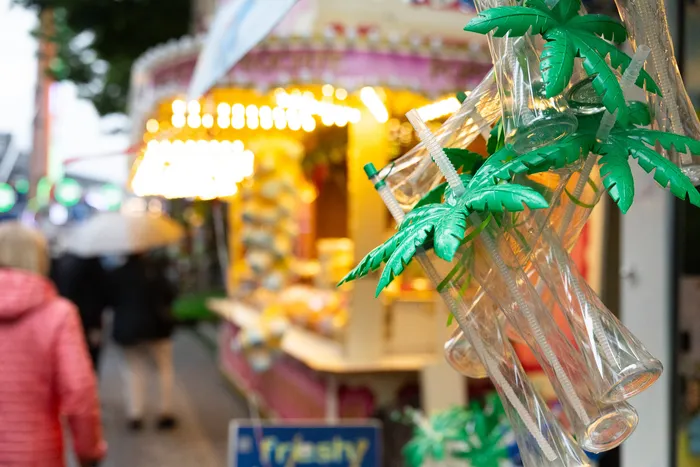 Dekoration aus Plastikpalmen und leeren Flaschen mit Strohhalmen vor einem Kirmesstand.