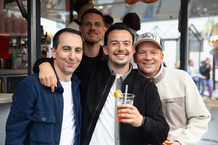 Vier Männer lächeln in die Kamera, einer hält ein Glas mit einem orangen Getränk. Im Hintergrund ist ein Kirmesstand zu sehen.