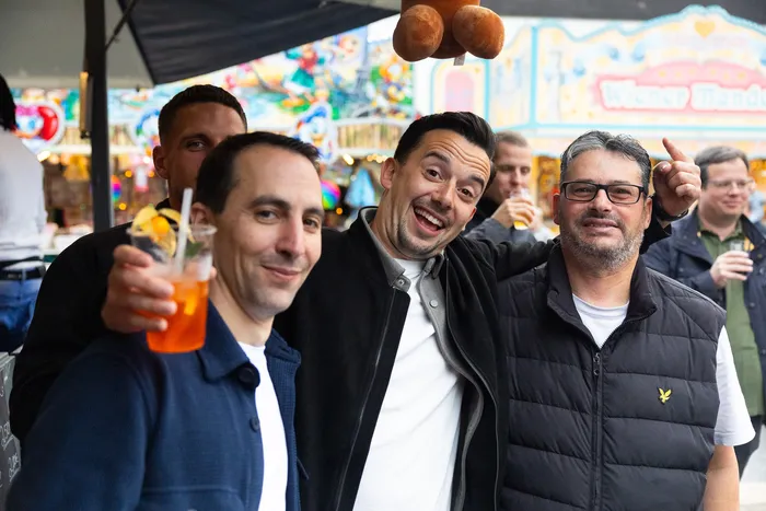 Drei Männer lächeln in die Kamera, im Hintergrund ein buntes Kirmesgelände mit Menschen und Buden.