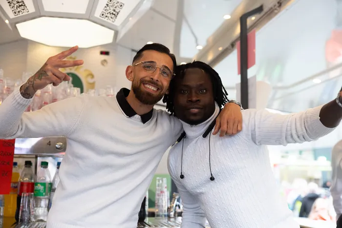 Zwei lächelnde Männer posieren eng umschlungen vor einer Bar mit Getränken im Hintergrund. Der Mann links trägt eine Brille und zeigt das Victory-Zeichen.