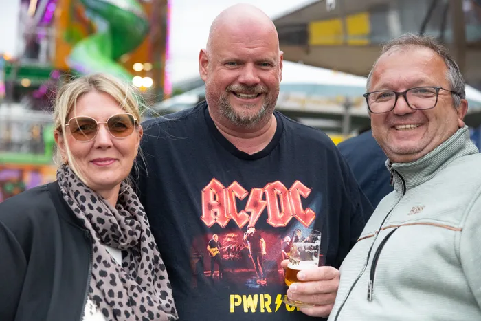 Drei lächelnde Menschen vor einer Kirmes. Der Mann in der Mitte trägt ein AC/DC-T-Shirt und hält ein Bierglas.