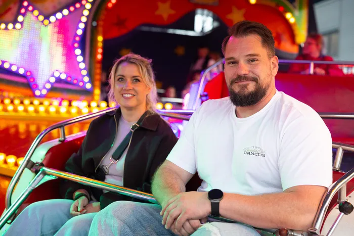 Zwei lächelnde Menschen sitzen in einem Fahrgeschäft auf der Kirmes, umgeben von bunten Lichtern und Dekorationen.