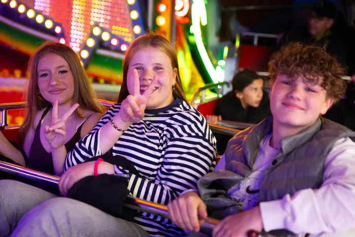Drei Jugendliche sitzen während der Kirmes in einer Fahrgeschäftsgondel und zeigen Victory-Zeichen.