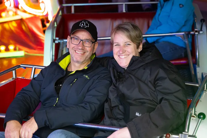 Lachendes Paar sitzt in einer Fahrgeschäftskabine, im Hintergrund bunte Lichter einer Kirmes.