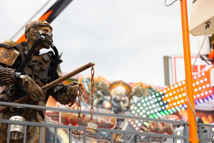 Eine detailreiche, menschengroße Figur in Skelettform mit mechanischen Elementen steht vor einem bunten Kirmes-Hintergrund.