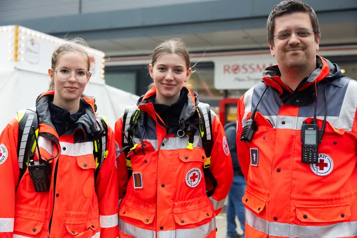 Drei ehrenamtliche Helfer des Deutschen Roten Kreuzes in orangefarbenen Warnwesten stehen vor einem Rettungswagen.