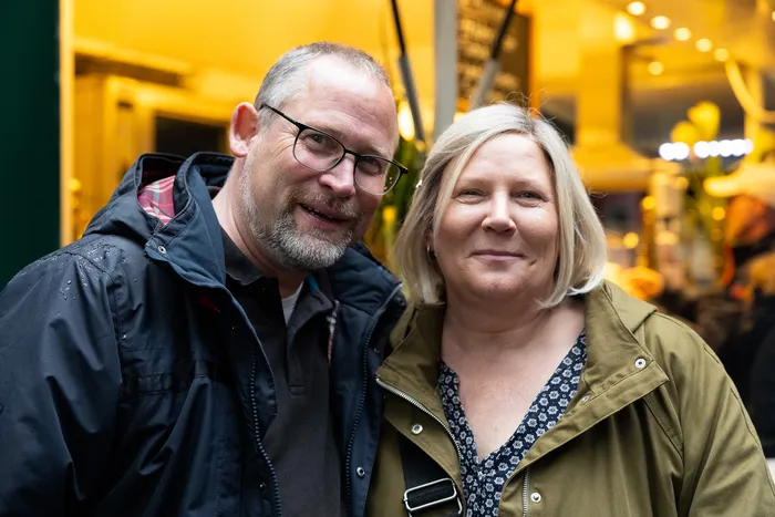 Ein Mann und eine Frau lächeln in die Kamera. Im Hintergrund ist ein Marktstand mit Blumen zu sehen.
