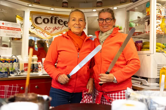 Zwei Frauen in roten Jacken halten jeweils ein Holzschwert und lächeln in die Kamera. Im Hintergrund ist ein Kiosk mit der Aufschrift „Coffee to go!“ zu sehen.