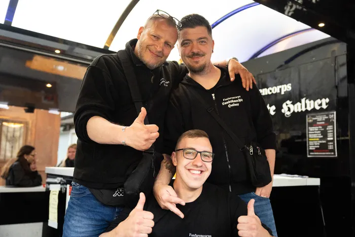 Drei Männer lächeln in die Kamera und zeigen Daumen hoch. Sie tragen schwarze Jacken mit dem Aufdruck „Fanfarencorps“.