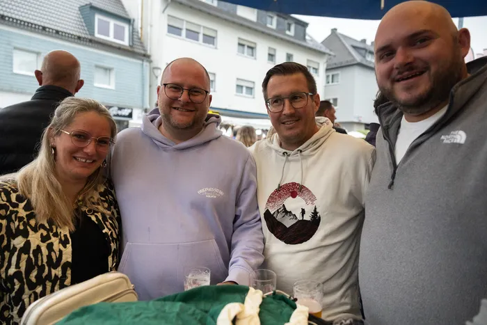 Vier lächelnde Menschen stehen eng zusammen vor einer Häuserkulisse. Sie tragen Freizeitkleidung und halten teilweise Getränke in der Hand.
