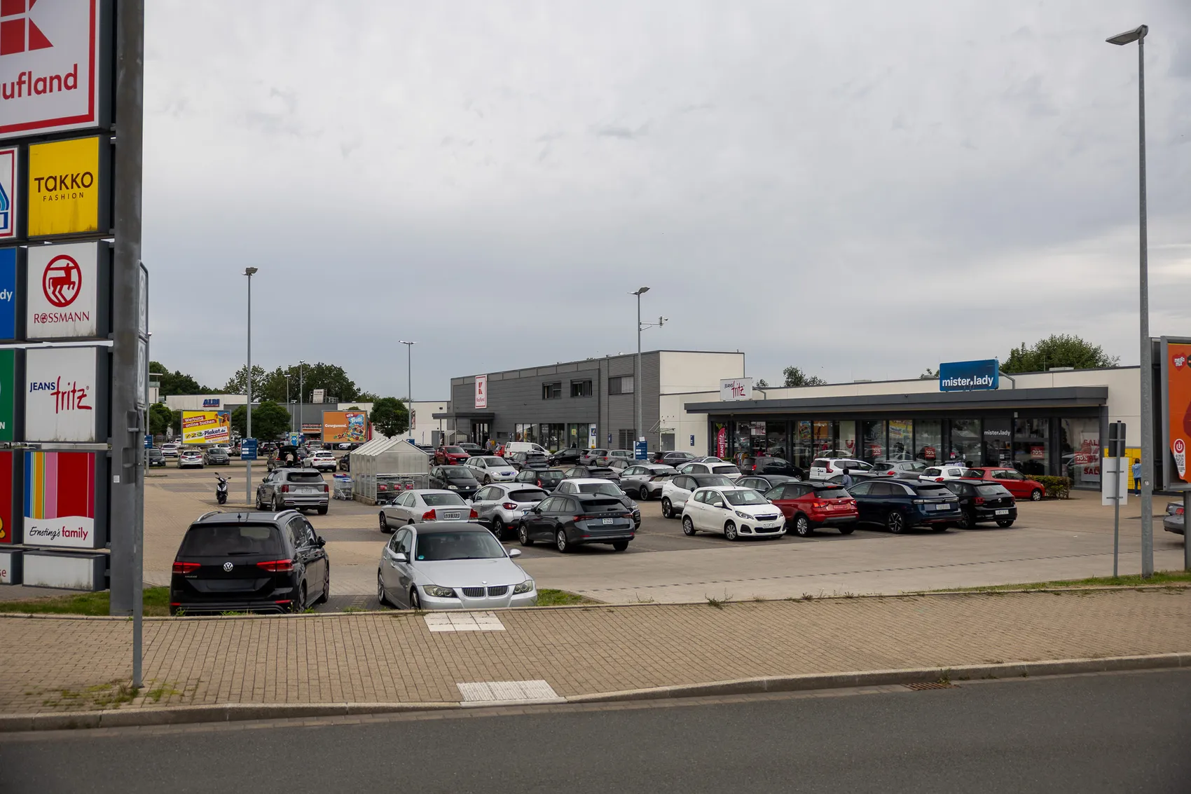 Ein Parkplatz mit zahlreichen Autos vor den Ladenfronten des Fachmarktzentrums Halver unter bewölktem Himmel.