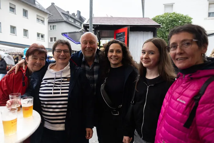 Sechs lächelnde Menschen stehen eng zusammen vor einer Stadtkulisse, einige halten Biergläser in der Hand.