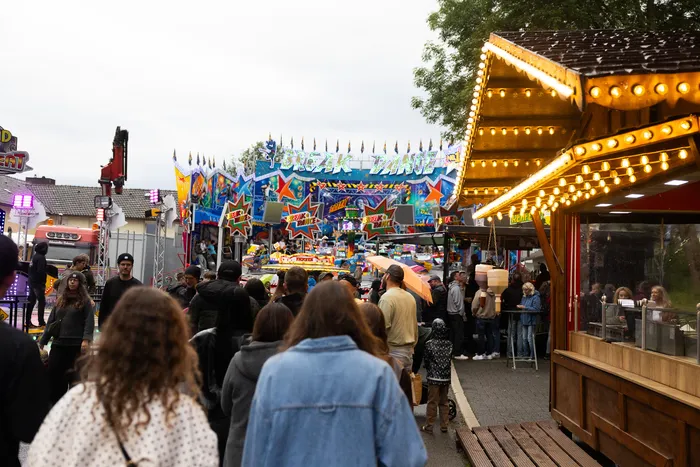 Menschenmengen bewegen sich auf einem Jahrmarkt zwischen beleuchteten Fahrgeschäften und Imbissständen.
