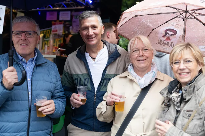 Vier Menschen stehen eng zusammen und halten Biergläser in der Hand, im Hintergrund ein Kirmesstand und ein Regenschirm mit einem bunten Muster.