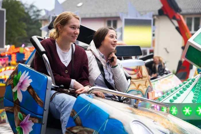 Zwei junge Frauen sitzen lächelnd in einer Karussell-Gondel, im Hintergrund weitere Fahrgeschäfte und Menschen auf der Kirmes.