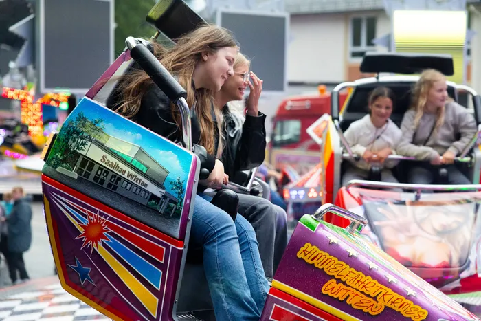 Zwei junge Frauen sitzen in einer pinkfarbenen Gondel einer Fahrgeschäftattraktion, während sie sich unterhalten. Im Hintergrund sind weitere Fahrgeschäfte und Besucher der Kirmes zu sehen.