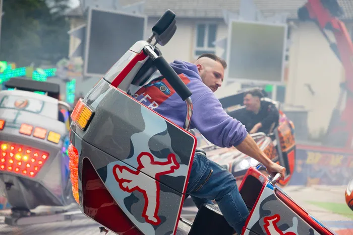 Ein junger Mann mit kurzgeschorenen Haaren lehnt sich in einer Karussell-Gondel nach außen, während die Anlage in Bewegung ist. Im Hintergrund sind weitere Fahrgeschäfte und Besucher zu sehen.