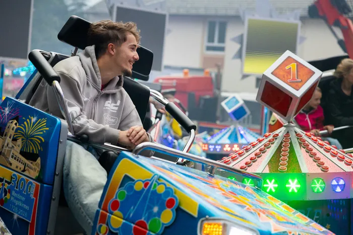 Ein junger Mann lächelt während der Fahrt mit einem Karussell auf der Kirmes. Im Hintergrund sind weitere bunt dekorierte Fahrgeschäfte zu sehen.