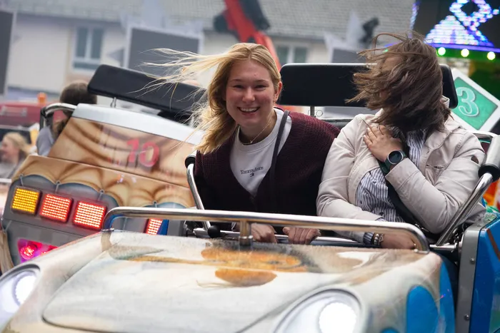 Zwei junge Frauen lachen während einer Fahrt auf einem bunt dekorierten Karussell. Im Hintergrund sind weitere Fahrgäste und Lichter zu sehen.